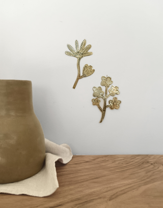 Notre duo de fleur graphique en laiton, accroché au mur, près d'un vase en Tadelakt couleur taupe qui est posé sur une planche en noyer et sur une serviette beige en coton